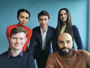 quantum dice team, five people, three are stood up and two sat down on chairs with a blue background.