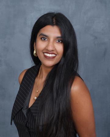 Sahana has long dark hair, and is smiling broadly at the camera. She wears a smart sleeveless top, and is pictured against a plain background.