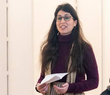 Dr Marina Pérez de Arcos stood giving a talk with a piece of paper in her hand, wearing a burgundy top and grey blouse. 