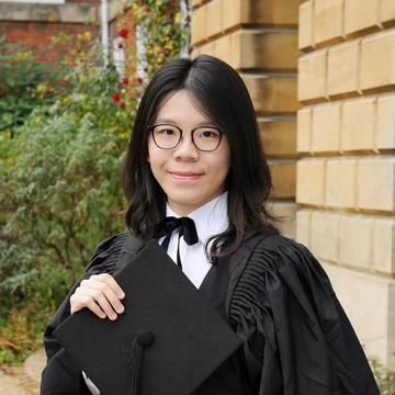 ceri ngai stood in full gown holding cap in front of a building.