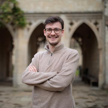 Francesc Puchal smiles towards the viewer, with arms folded. He stands in front of the St Cross arches. He has short brown hair and glasses.