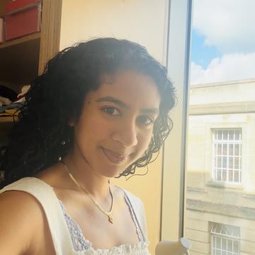 Ashley Duraiswamy has dark curly hair. She wears a white top and a gold necklace and smiles towards the viewer.