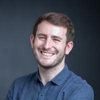 alex caro head and shoulders shot, smiling with a dark blue open shirt with a dark background.