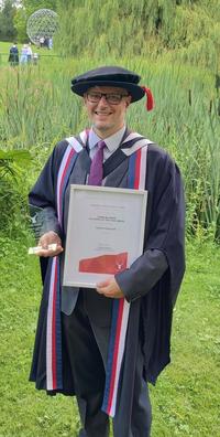 Dr Lorenzo Santorelli stood in garden wearing full gown, holding framed certificate and trophy.