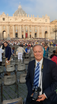 Peter Nockles stood in front of a historic building, wearing a full suit, and camera hanging from his neck.