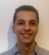 Head shot of Youssuf Saleh smiling with open collar shirt on white background.